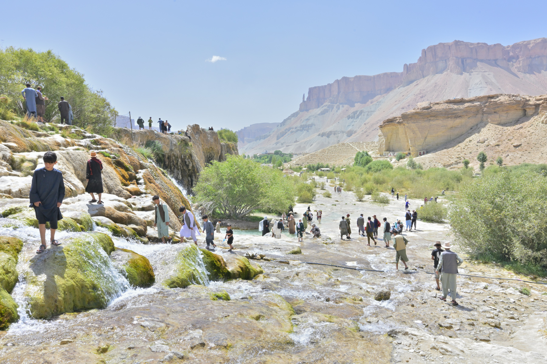 Dozens of people wade and walk across a wide travertine shelf where turquoise water spills in shallow terraces, pale cliffs and a dry valley stretching behind them.