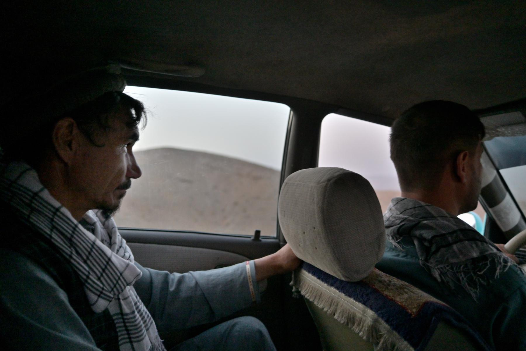 Two silhouetted men seen from the back seat of a moving car, a hazy pale ridge of desert visible through the dust-filmed side window.