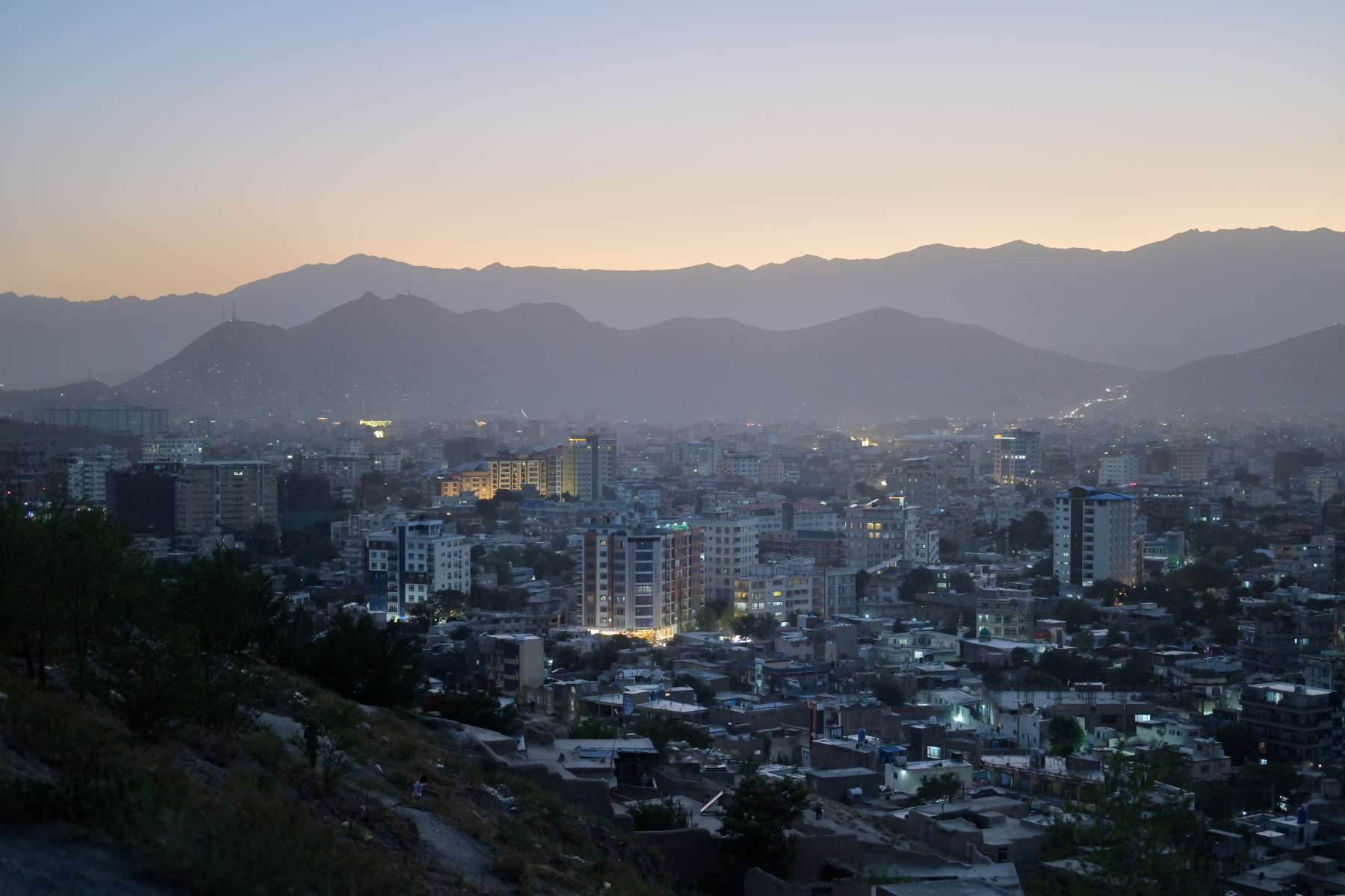 A dense hillside city spreads across a valley floor at dusk, scattered windows beginning to glow amber, mountains fading to silhouette against a darkening sky.
