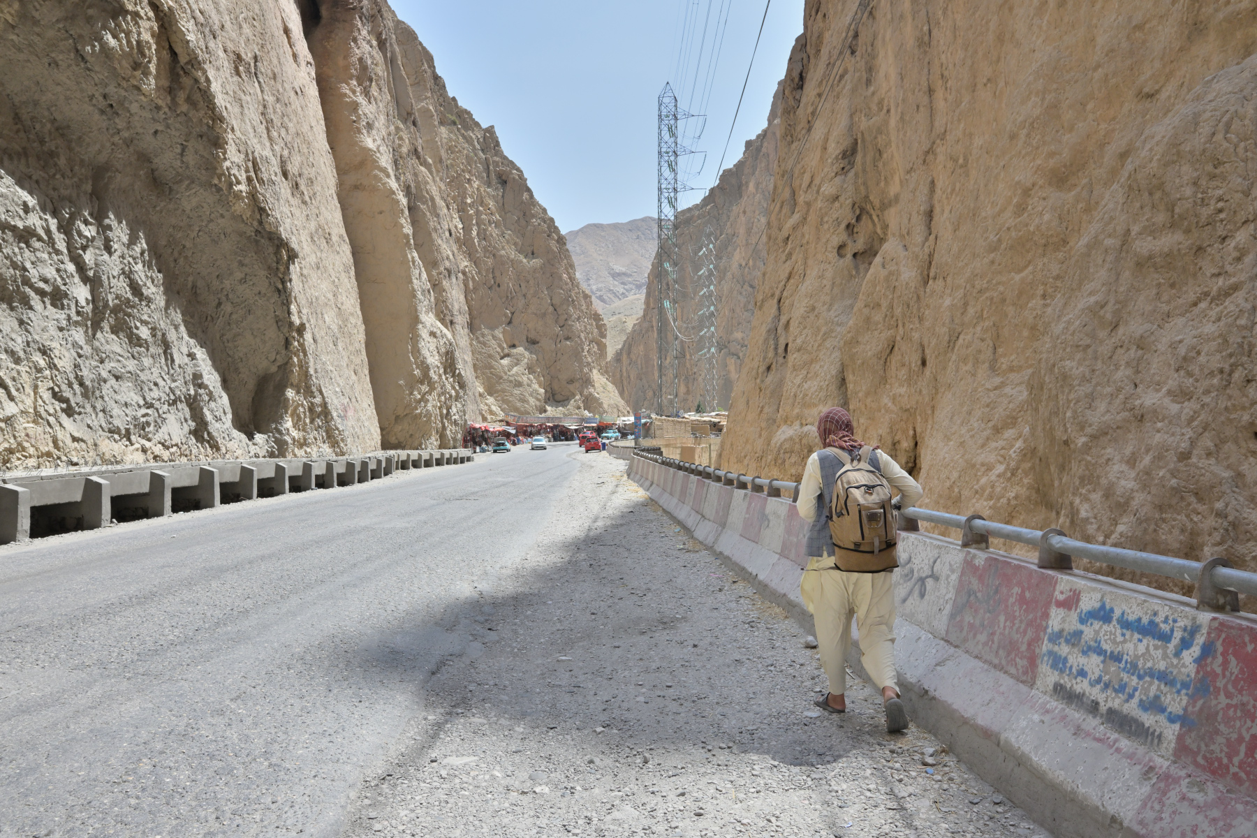 An empty road cutting through a steep rock gorge, a man walking away from the camera toward a distant market.