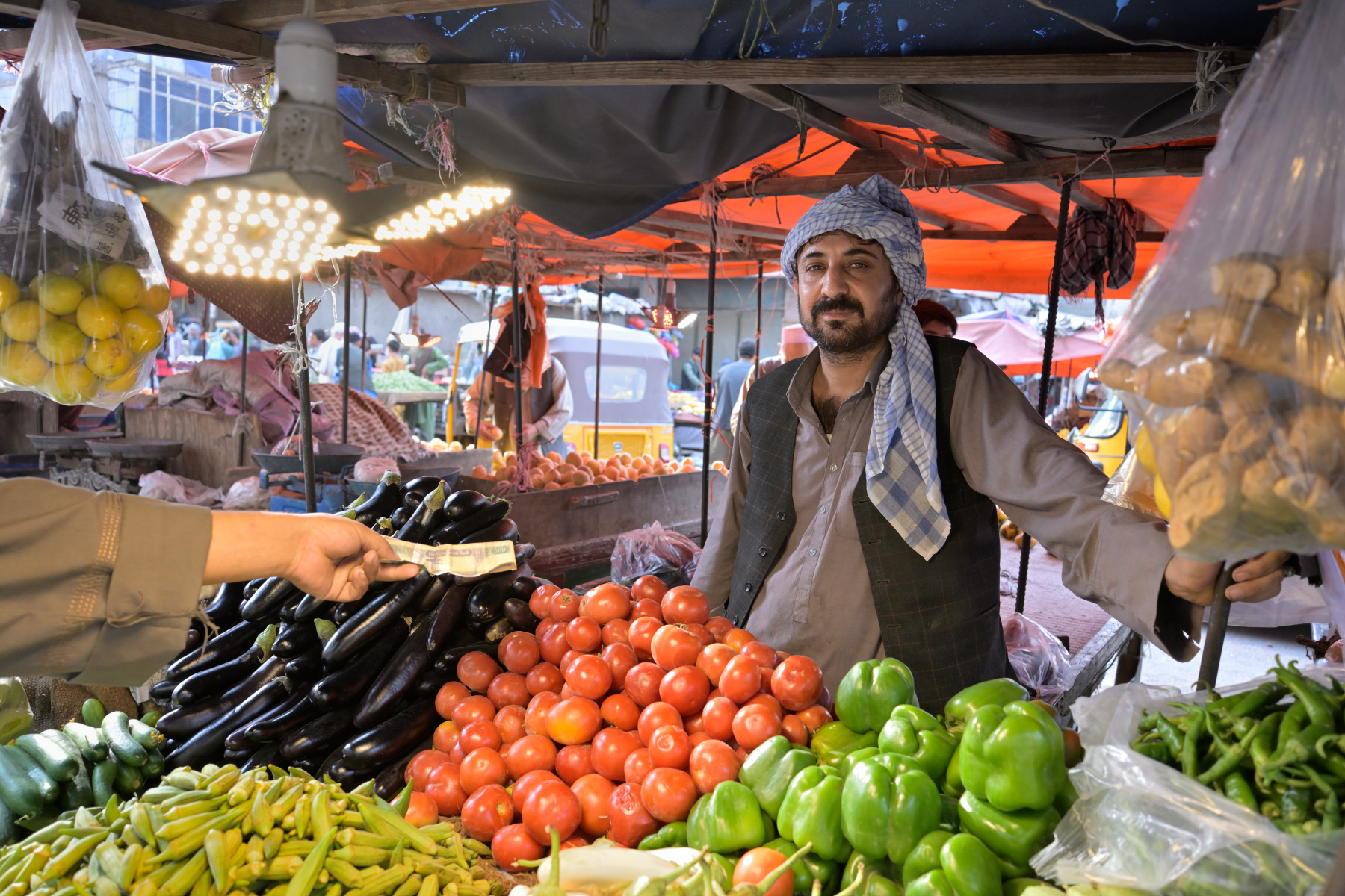 A market vendor in a checked headscarf stands behind a stall piled with tomatoes, eggplant, peppers and okra, a customer's hand passing money across the produce.