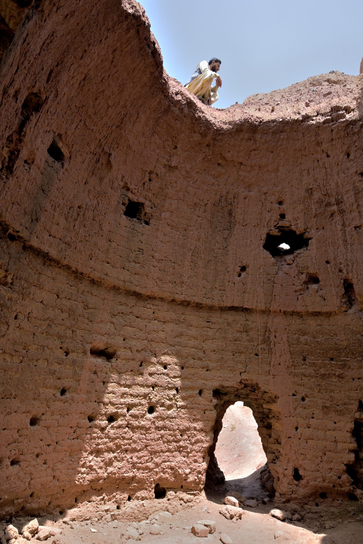 An adobe wall riddled with small carved niches; a man peers over the top edge, half in shadow.