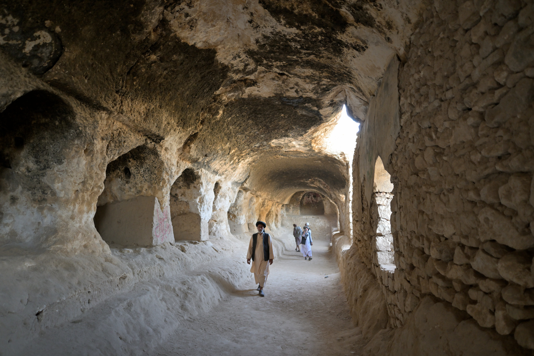 A vaulted stone corridor carved from pale rock, a man walking toward the camera, two more figures further back near a bright opening in the wall.