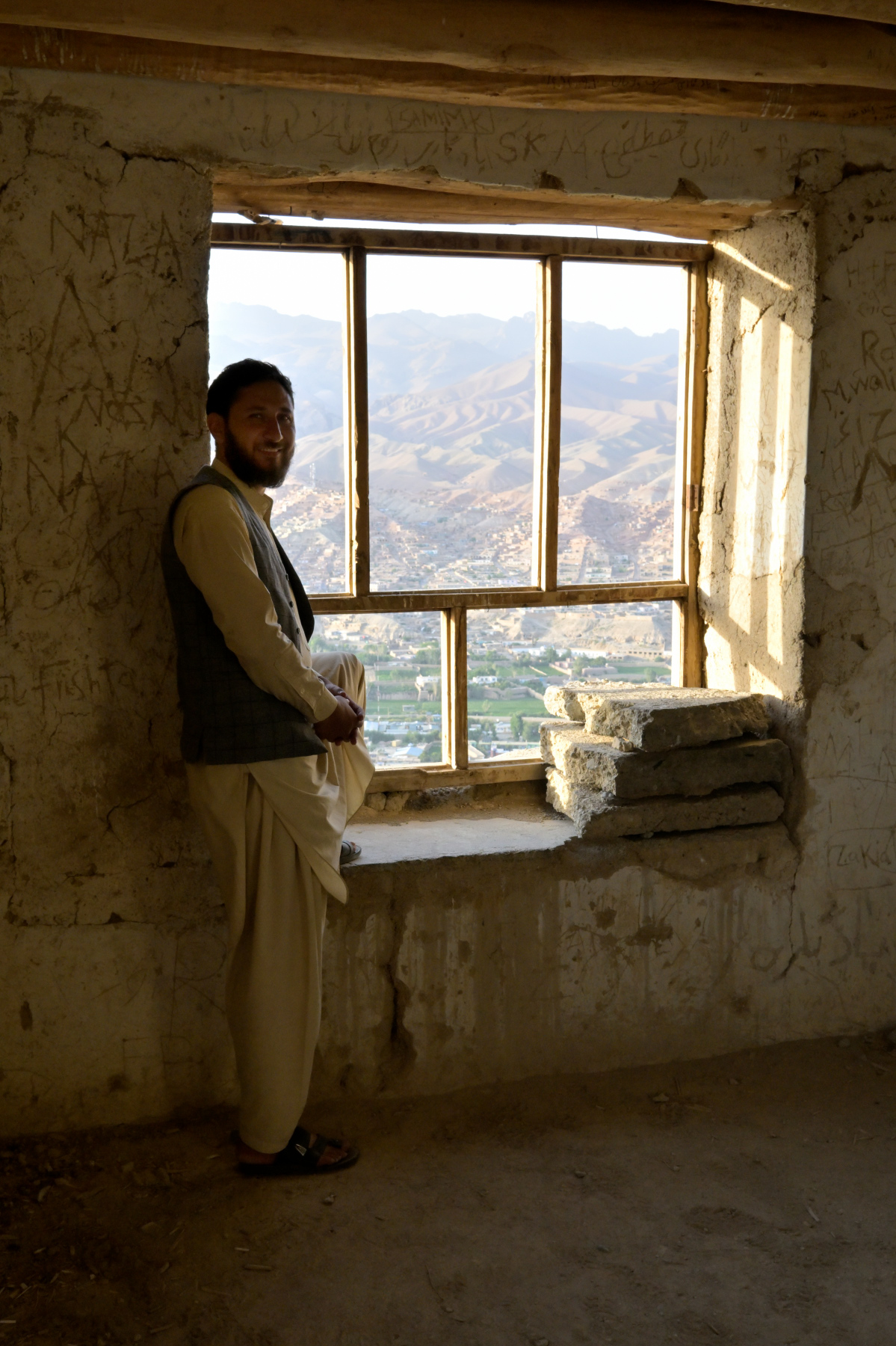 A man leaning by a ruined window opening, framed against layered mountains and a hillside town.